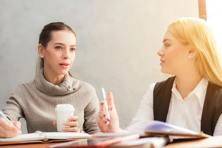 Two female office workers talk as they review reports open on the table before them.