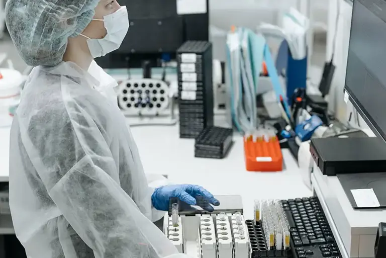 Masked scientist in a lab stands in front of a computer, while they check a large batch of vials