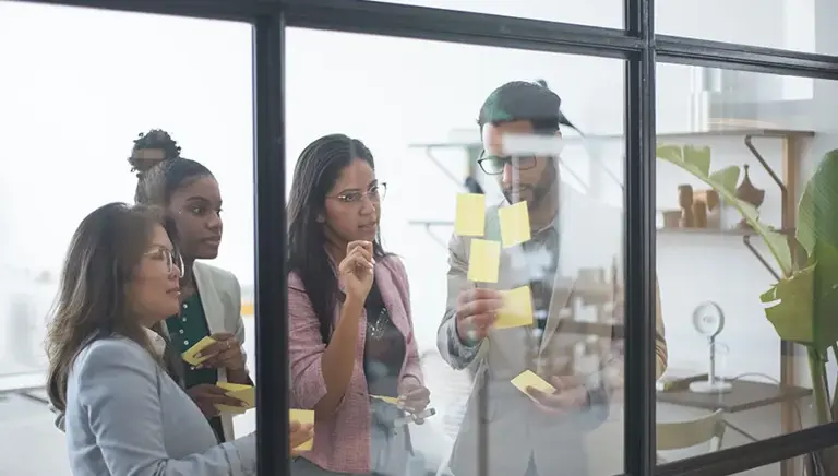 Four office workers are seen through a window into a conference room where they are working on a series of post-it notes on the glass.