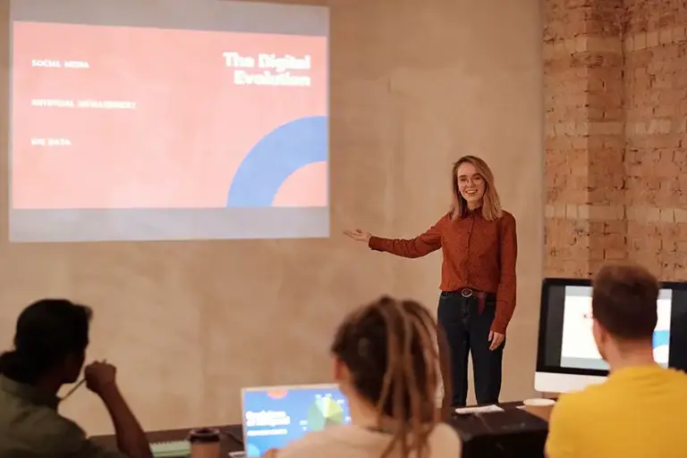 Presenter stands in room in front of her projected presentation, facing the audience and talking