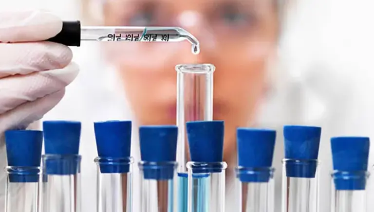 Close up of a rack of test tubes with a scientist adding a single drop from a pipette into the middle test tube.