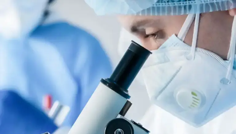 Close up side view of a masked scientist looking down a microscope