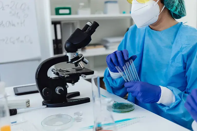 Masked scientist, in lab sat at a microscope holding a set of vials