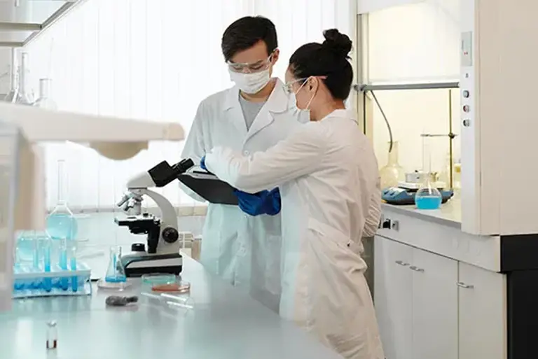 Two scientists discussing are standing in a lab in front of a microscope on the bench.