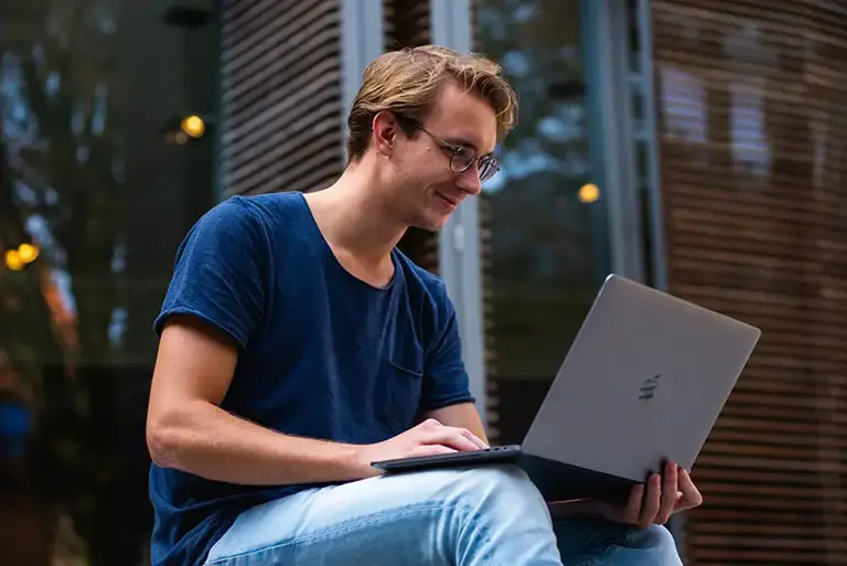 Casually dressed young man, sits outside, working on a laptop that rests on his knees