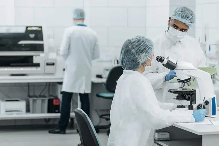 Three scientists working in a lab in conversation while one looks through a microscope