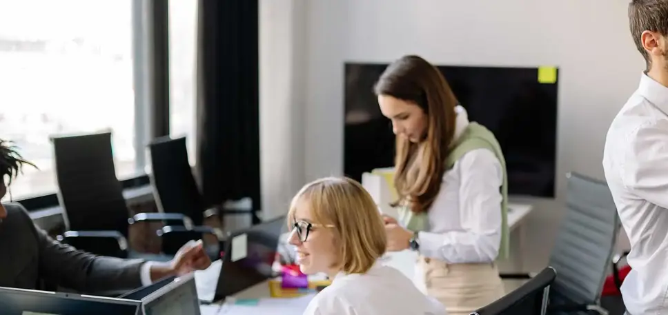 Two team members working at a long office table while another stands and writes a post it note on the wall