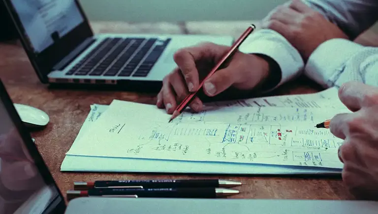 Close-up of the hands of two office workers writing notes on a desk between to open laptops